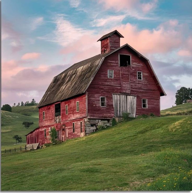 an old red barn sits on the side of a grassy hill with a pink sky in the background