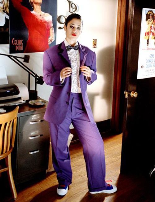 a man in a purple suit and bow tie standing next to a poster on the wall