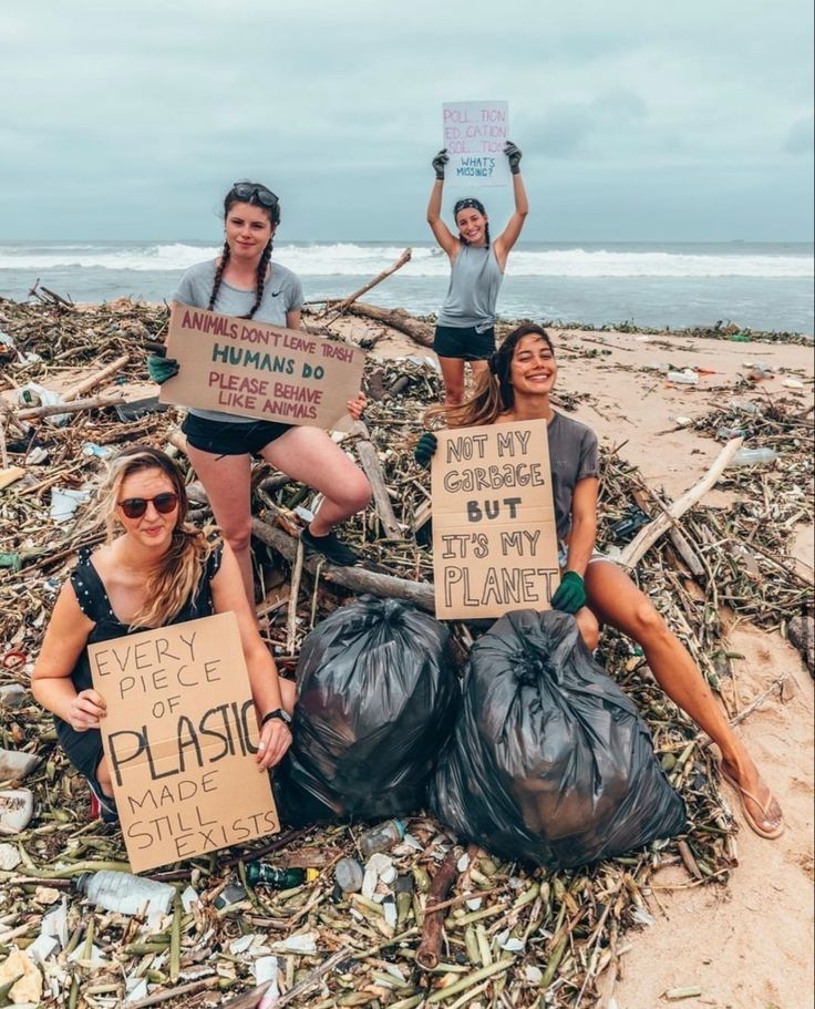 three women holding signs on the beach with trash bags and garbage in front of them