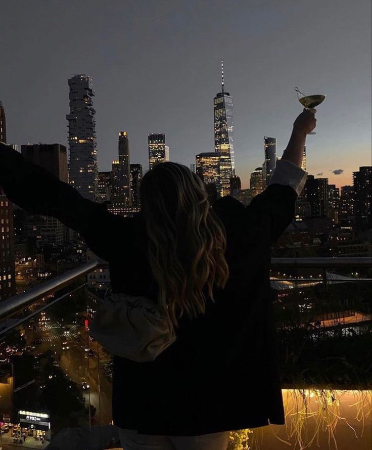 a woman standing on top of a building holding a wine glass in her hand and looking at the city