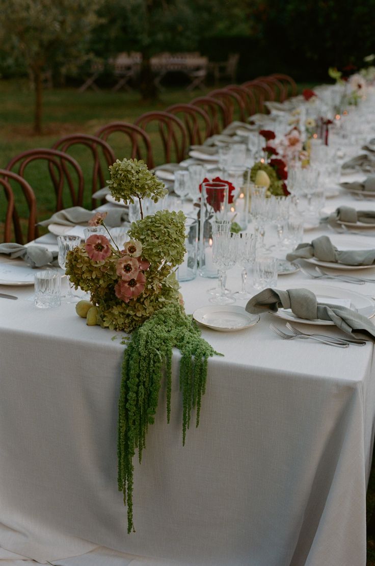 a long table is set with white linens and place settings for an outdoor dinner