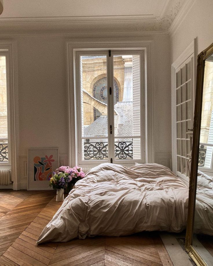 a bed sitting in front of two windows next to a mirror on top of a wooden floor