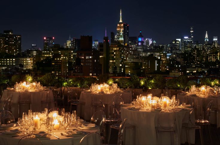 an outdoor dining area with tables and chairs set up in front of the city skyline at night