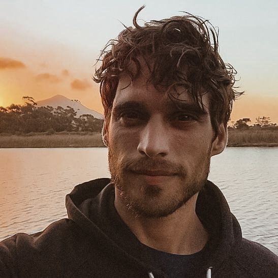 a man standing in front of a body of water with the sun setting behind him