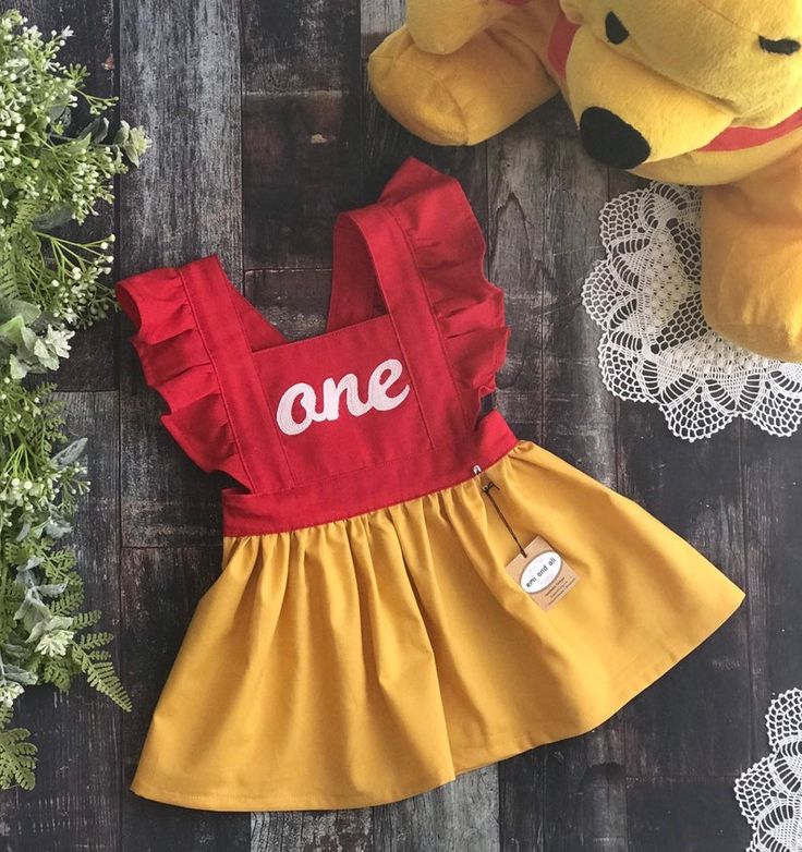 a teddy bear sitting next to a red and yellow dress with the word one on it