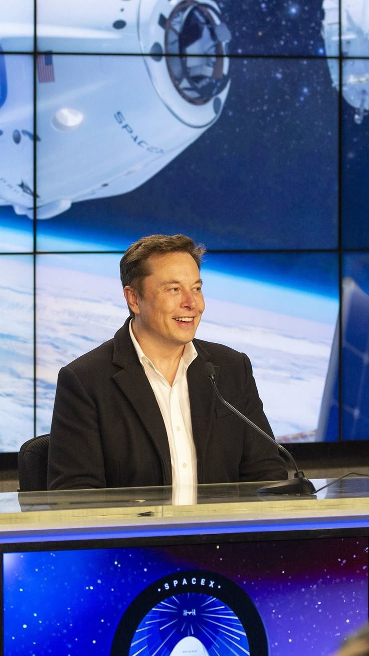 a man sitting at a podium in front of a television screen with the space shuttle behind him