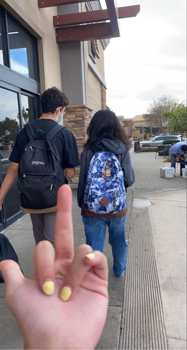 two people walking down the sidewalk with backpacks on their backs and one holding up a peace sign