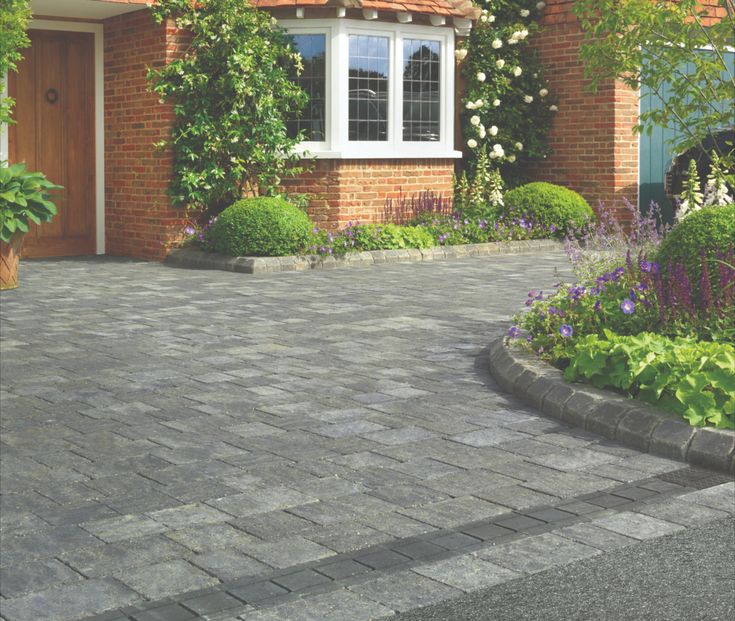 a brick driveway in front of a house with flowers and shrubs around it, along with a potted plant on the side