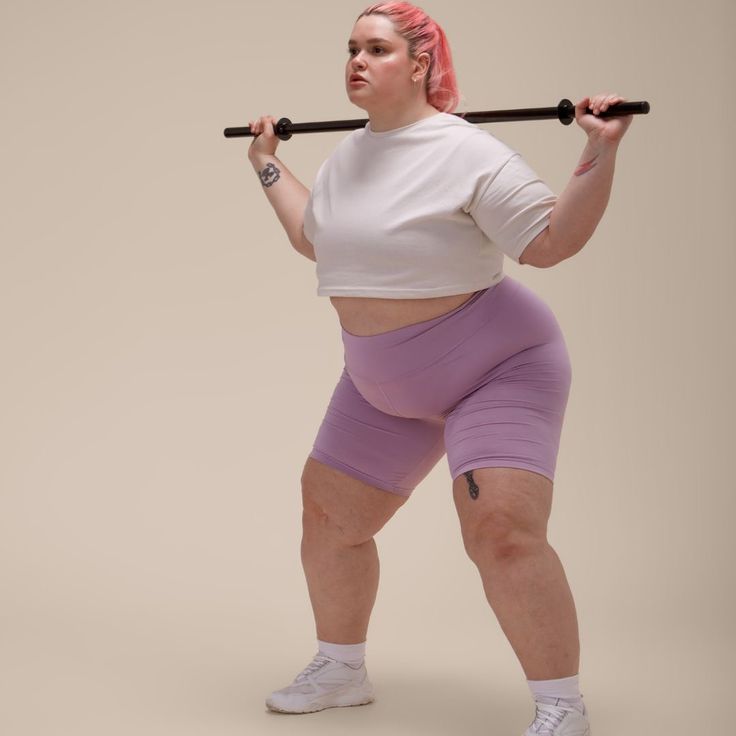 a woman with pink hair holding a barbell in her right hand and wearing purple shorts