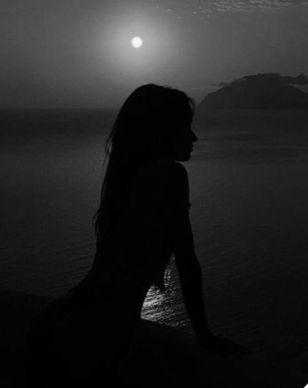 a woman sitting on the edge of a cliff looking out to sea at night time