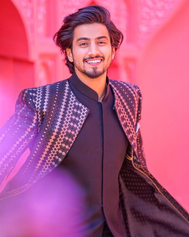 a man with long hair wearing a black shirt and jacket smiling at the camera while standing in front of a pink background