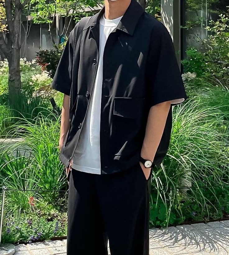 a man standing in front of some bushes and trees wearing a black shirt with his hands in his pockets