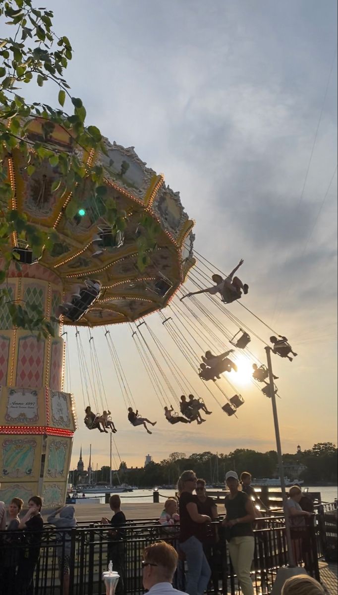 people are gathered around a carnival ride at the park as the sun sets over the water