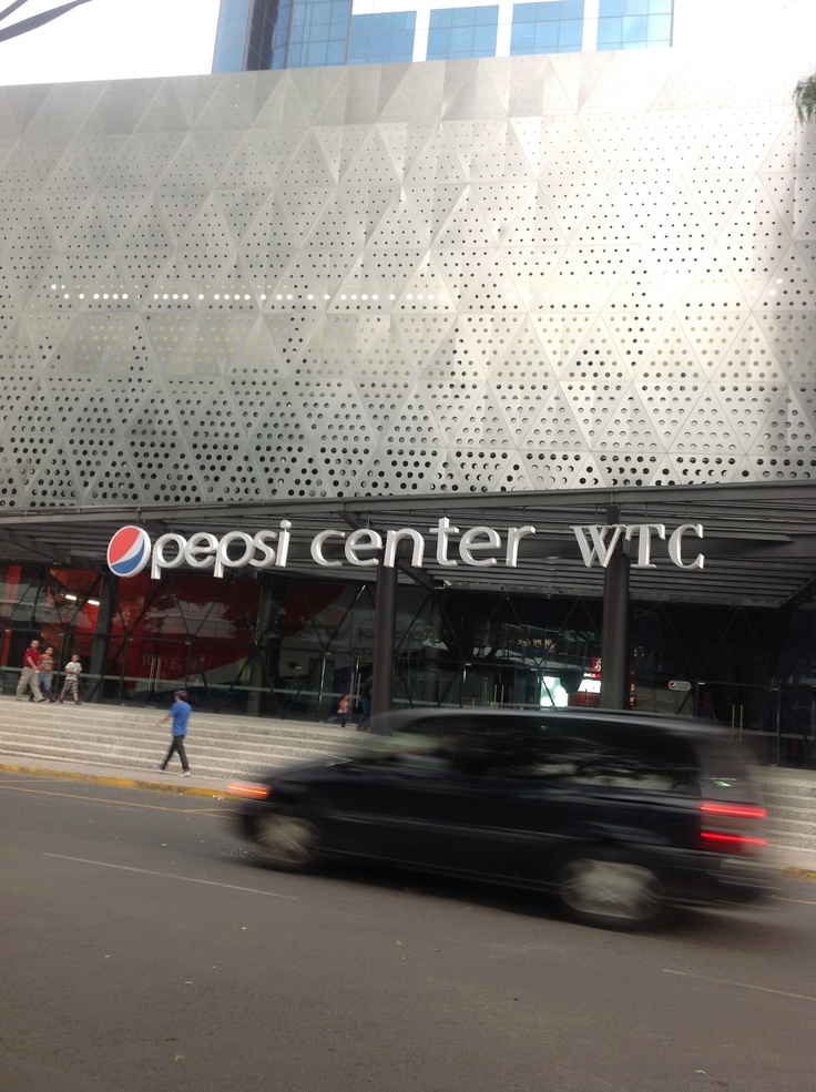 a black car driving past a pepsi center with people walking on the sidewalk and cars passing by