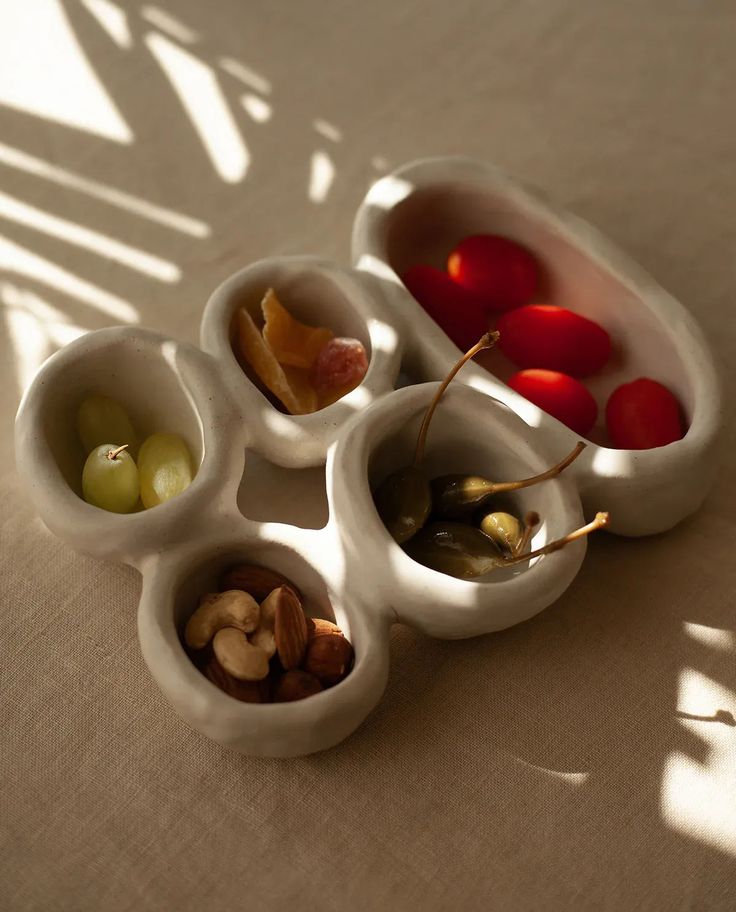 four white bowls filled with different types of fruit and nuts on a tablecloth covered surface