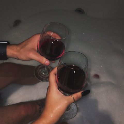 two people toasting with red wine in their hands on an ice - filled bathtub