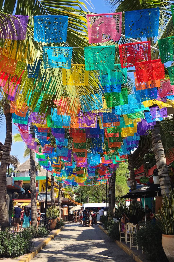 many colorful umbrellas hanging from the side of a road next to palm trees and buildings