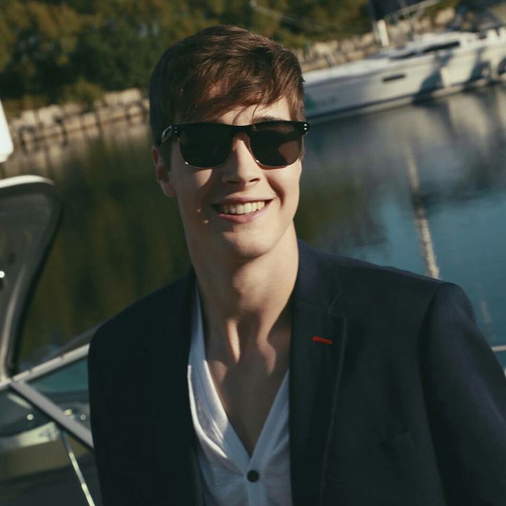 a young man wearing sunglasses standing next to a boat in the water at a marina