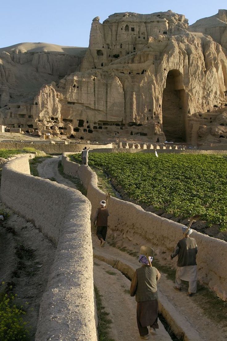 some people are walking down a path in front of an old rock formation and mountains