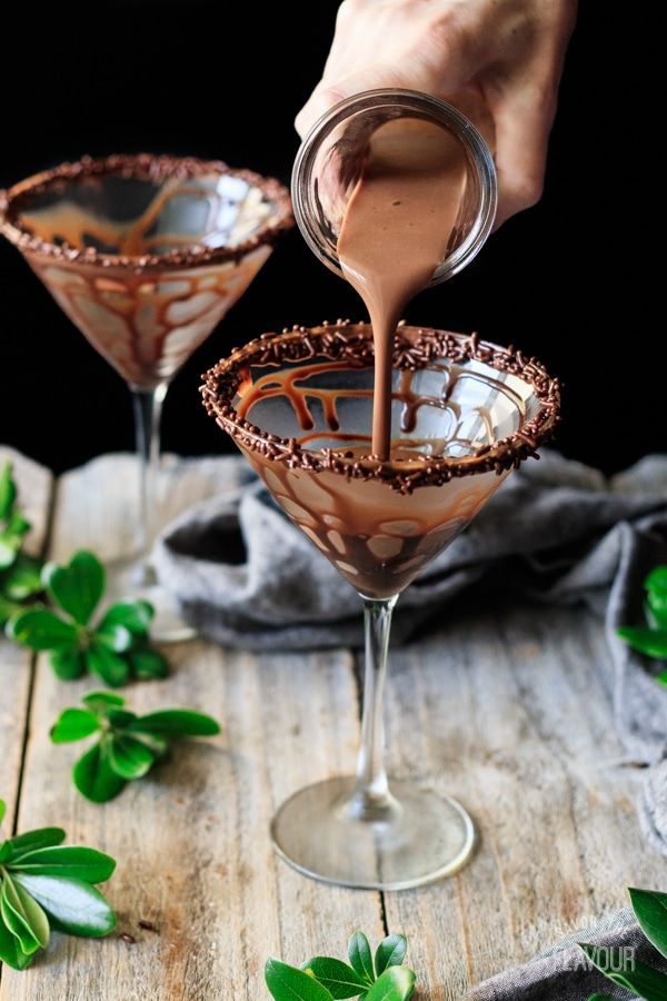 someone pouring chocolate into two martini glasses on top of a wooden table with green leaves