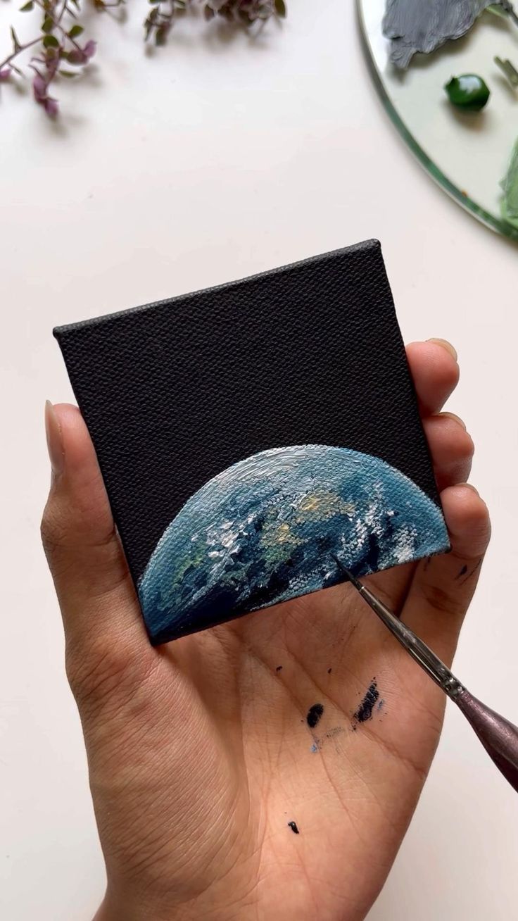 a hand holding a piece of black paper with the earth painted on it and flowers in the background