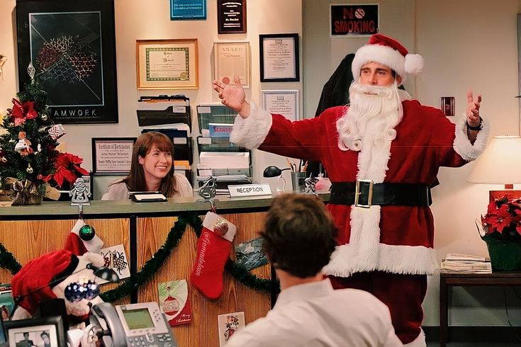 a man dressed as santa clause standing in front of a desk with people around him