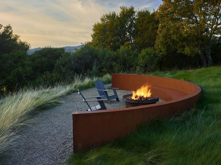 a fire pit in the middle of a grassy area with two chairs around it and trees in the background