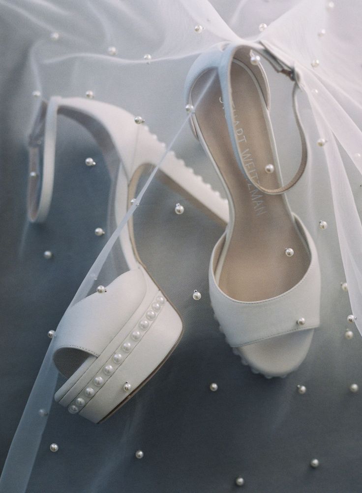 a pair of white wedding shoes with pearls on the heel and veil in the background