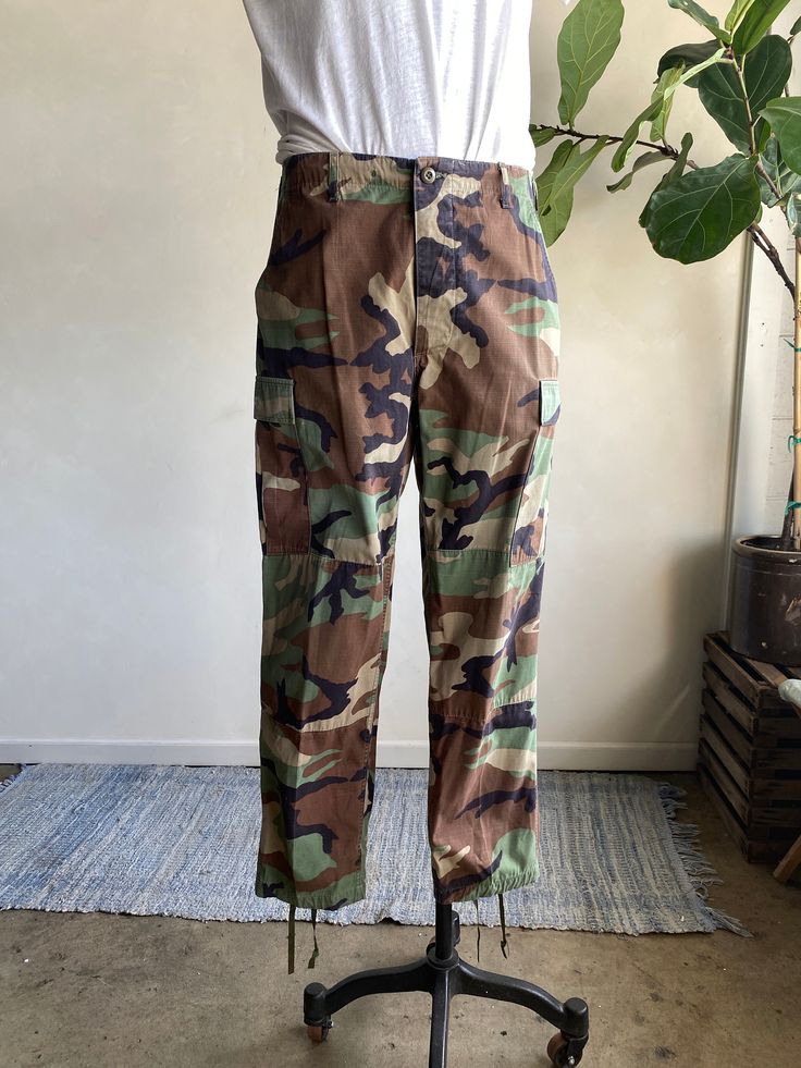 "Vintage Military Woodland Camo Button Fly Poplin Cargo Pants. Military issued pants with a camo pattern made from a cotton/nylon fiber blend. Pants have a button fly closure and adjustable straps at waist. Oversized cargo side pockets and straps and bottom hems. Condition overall is great with no major flaws noted. No rips, tears, or staining. Please see all photos for details. Refer to measurements below to ensure a proper fit  16.5\" across waist (size 33) 18\" across hip 31\" inseam" Camouflage Utility Cargo Pants With Belt Loops, Outdoor Camouflage Cargo Pants With Patch Pockets, Military Style Camouflage Cargo Pants, Military Camouflage Cargo Pants With Patch Pockets, Camouflage Cotton Parachute Pants With Side Pockets, Cotton Camouflage Parachute Pants With Side Pockets, Khaki Cargo Pants For Hunting With Pockets, Full Length Camouflage Cotton Cargo Pants, Khaki Cargo Pants With Pockets For Hunting