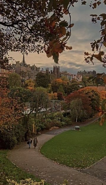 two people walking down a path through a park with trees and buildings in the background