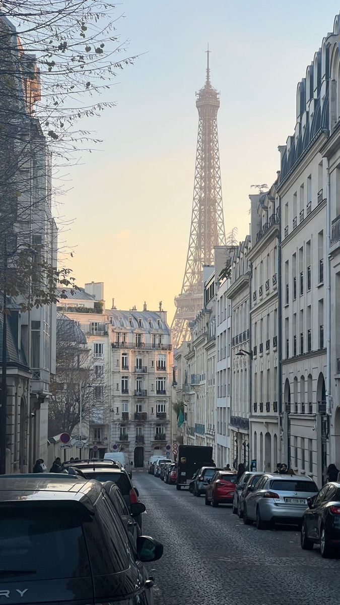 the eiffel tower is in the background of this street lined with parked cars