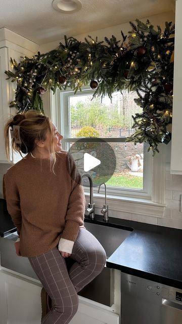 a woman sitting on top of a kitchen counter next to a window covered in christmas wreaths