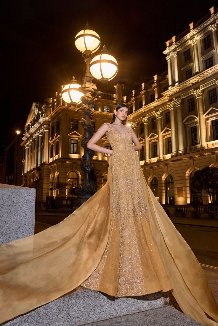 This golden gown set features embroidery in delicate gold coloured pearls in an abstract pattern. The plunging neckline of the gown has intricate cut work. The shapely gown also shows panels with heavy shrinkingly. An exaggerated satin organza bow has been used for styling purposes.From Seema Gujral's For the love of London collection. DELIVERY TIMEPlease allow 8-12 weeks for your outfit to arrive. FABRIC DETAILSNet Professional cleaning only. Gold V-neck Evening Dress With Sweep Train, Luxury Floor-length Gown With Sheer Bodice, Luxury Champagne Gala Gown, Formal Gown With Sheer Bodice And Long Train, Glamorous Evening Dress With Long Train, Luxury Evening Gown With Long Train, Floor-length Organza Evening Dress With Detachable Train, Gold V-neck Evening Dress For Gala, Silk Ball Gown For Gala