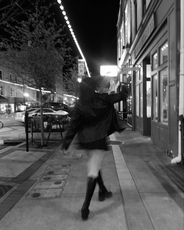 a woman walking down the sidewalk in front of a building at night with lights on