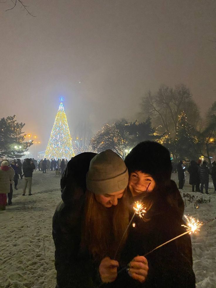 two people holding sparklers in front of a lit up christmas tree on a snowy day