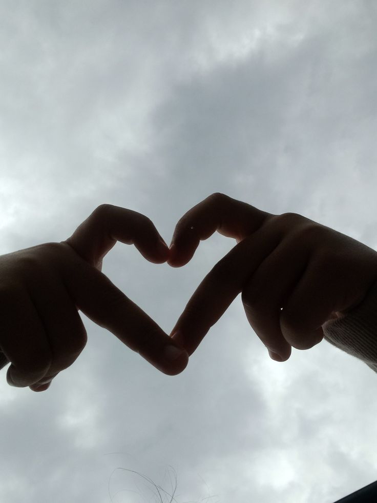 two hands making a heart shape with their fingers in front of a cloudy blue sky