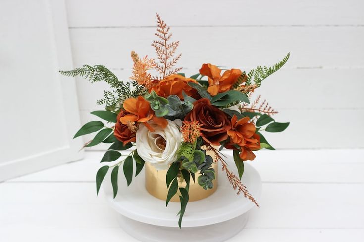 an arrangement of flowers in a gold vase on a white pedestal with greenery and foliage