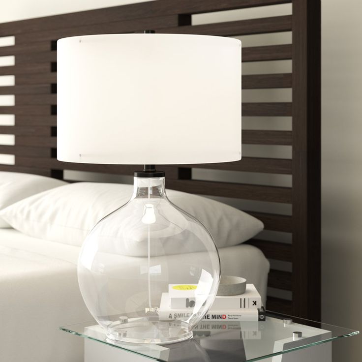 a glass table with a lamp on top of it next to a white pillow and bed