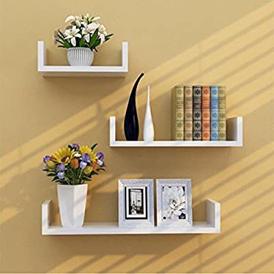 two white shelves with vases and flowers on them against a tan wall, next to bookshelves