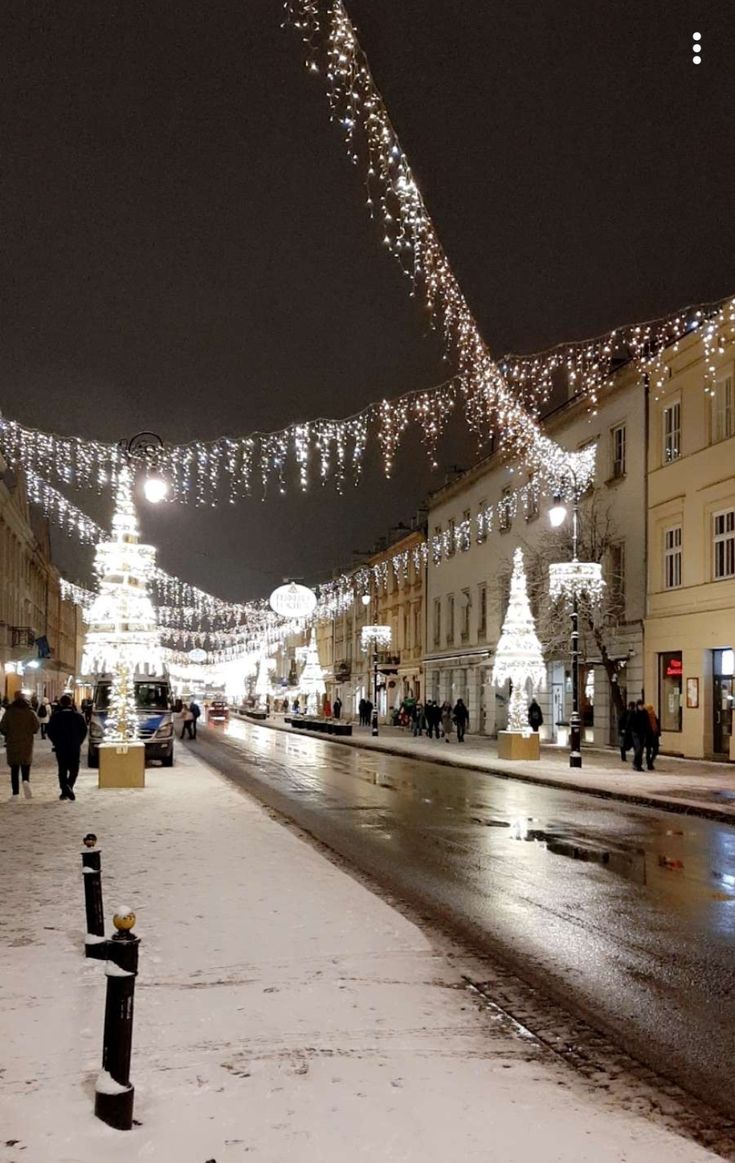 people walking down a snowy street with christmas lights on the buildings and cars parked in front of them