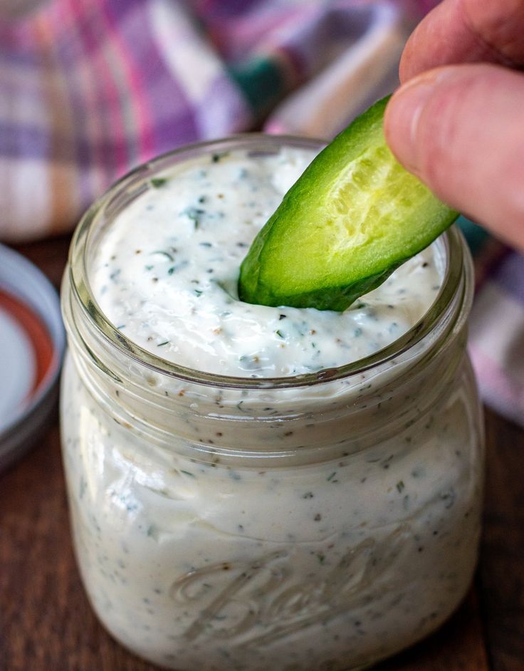 a person dipping a cucumber into a jar of ranch dressing on a wooden table