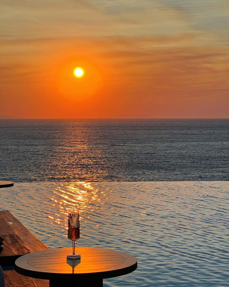 the sun is setting over an ocean with two wine glasses on a table next to it