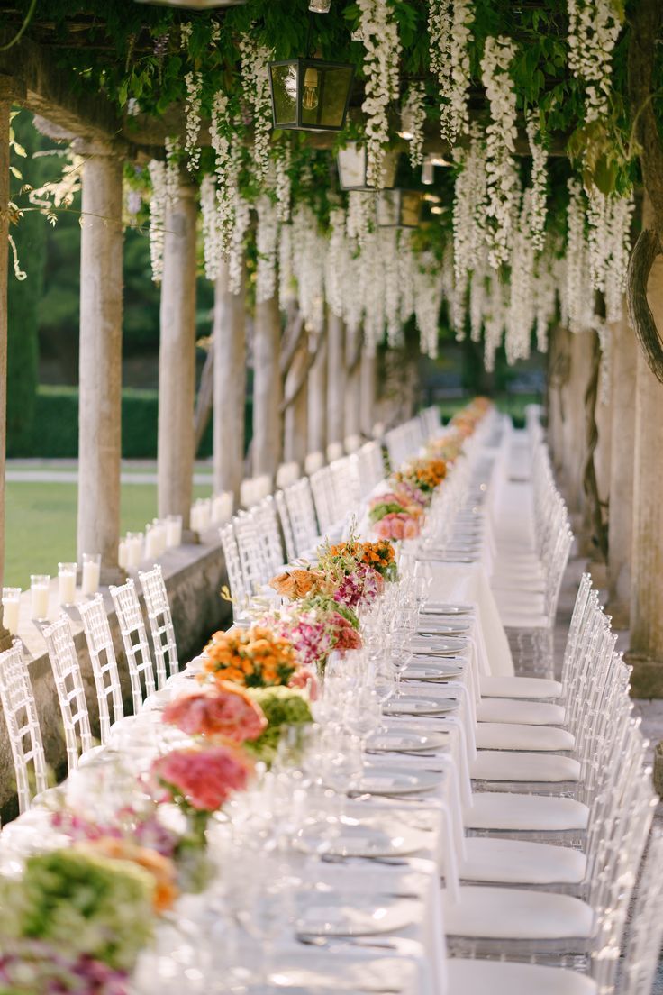 a long table is set with white chairs and flowers hanging from the pergolated roof