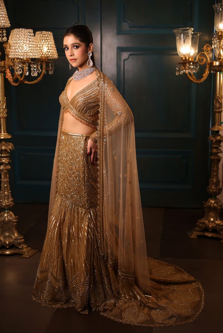 Metallic gold fishcut lehenga with all-over sequin, crystal, and cutdana embroidery with a flowing trail at the back. Paired with a delicate full sleeves blouse with hanging tassel detailing and embroidered dupatta in net.DELIVERY TIMEPlease allow 8-12 weeks for your outfit to arrive.FABRIC DETAILSNetProfessional cleaning only. Anarkali Style Hand Embellished Pre-draped Saree, Festive Embellished Pre-draped Saree With Long Sleeves, Festive Embellished Long Sleeve Pre-draped Saree, Festive Long Sleeve Embellished Pre-draped Saree, Traditional Pre-draped Saree With Sheer Dupatta And Long Sleeves, Long Sleeve Sharara With Dupatta For Reception, Gold Sharara With Sequins In Traditional Drape, Designer Long Sleeve Lehenga With Sheer Dupatta, Traditional Drape Gold Sharara With Sequins