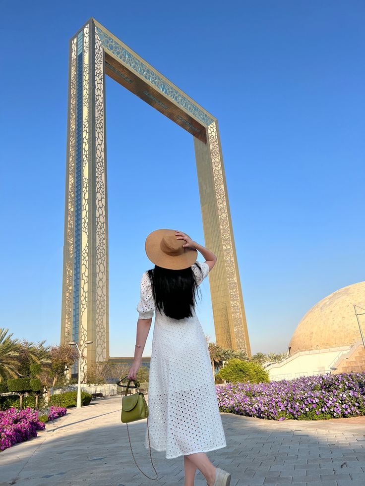 a woman in a white dress and straw hat walking towards a large arch with purple flowers