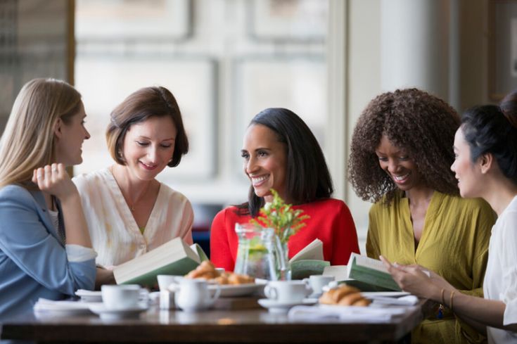 four women sitting at a table with books and coffee in front of them, talking