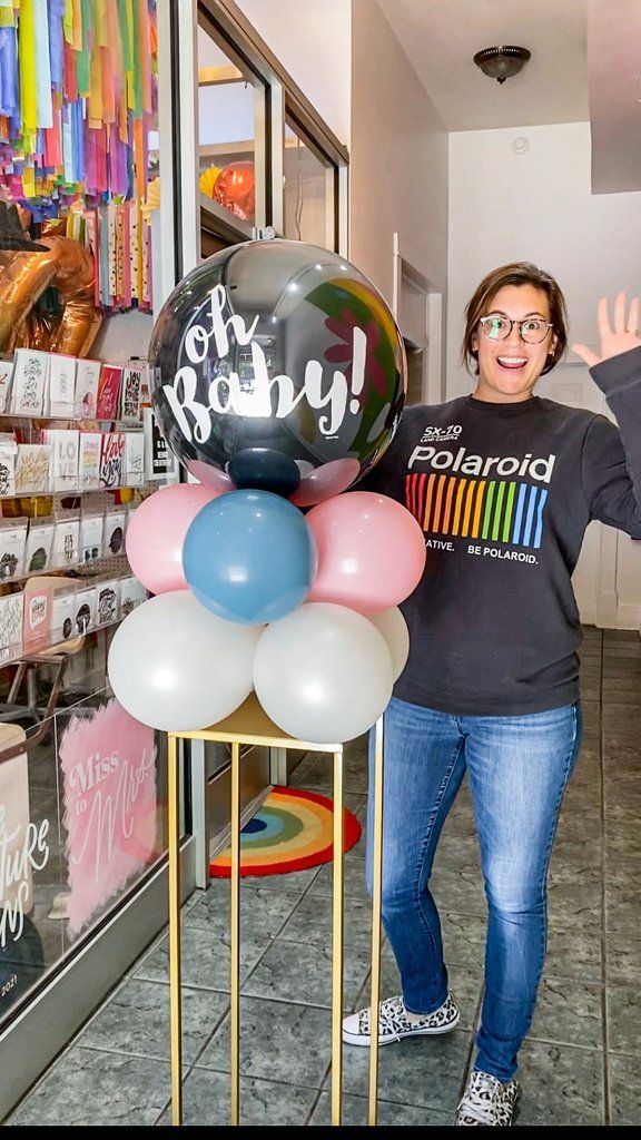 a woman standing in front of a store with balloons on the counter and some signs behind her