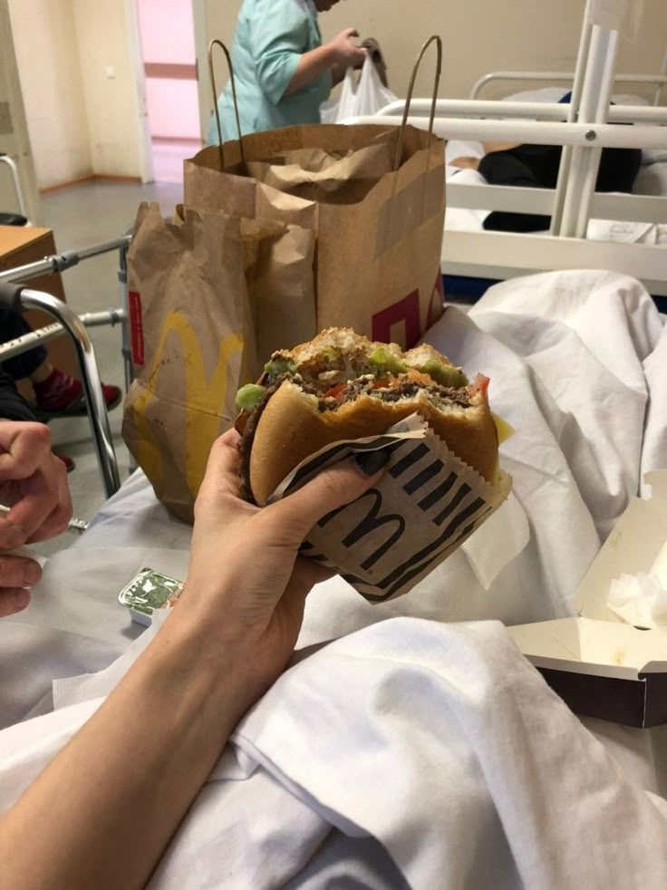 a person holding a sandwich in their hand while sitting on a hospital bed with other people nearby