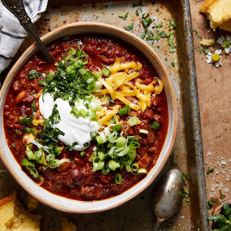 a bowl of chili with sour cream on top and bread in the background next to it