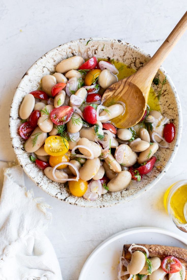 a white bowl filled with lots of food next to a wooden spoon on top of a plate